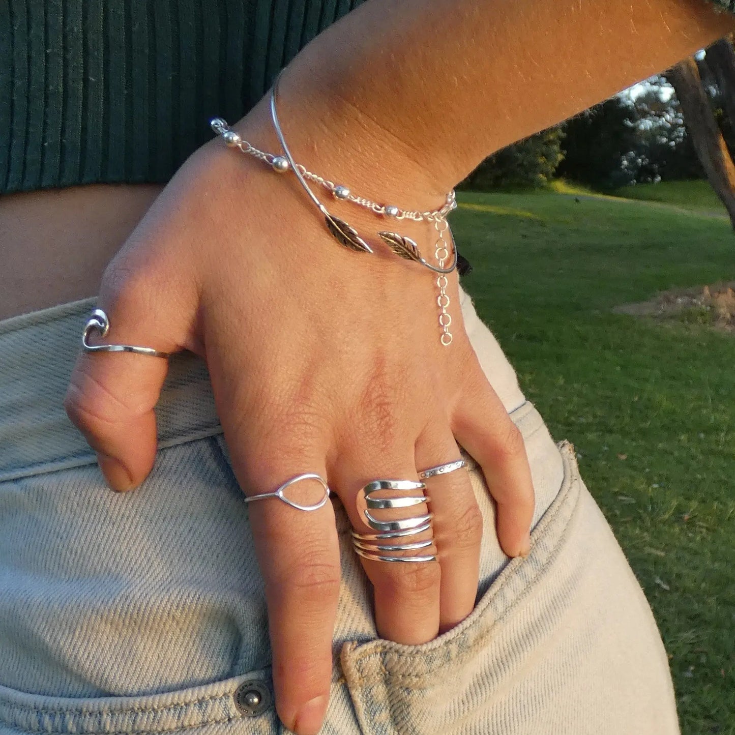 Close-up of a hand wearing silver statement rings, a feather bangle and bead bracelet against a natural background